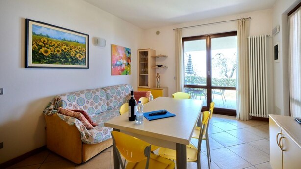 Un comedor con una mesa, sillas y un sofá. Hay un cuadro en la pared, un radiador y una ventana con cortinas.