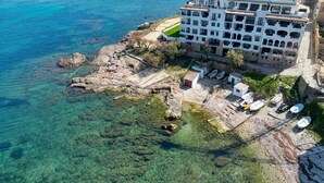 Vista aerea di un hotel sulla costa con diversi piani, una spiaggia rocciosa e acque blu cristalline.