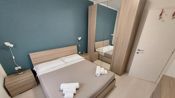 Un lit bien fait, avec du linge de lit blanc et des serviettes pliées, une table de chevet avec une lampe et un miroir qui reflète la pièce.