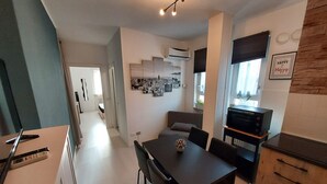 Une cuisine moderne dotée d’un four encastré, un coin repas avec une table et des chaises en bois foncé, et un salon avec un canapé et un téléviseur.