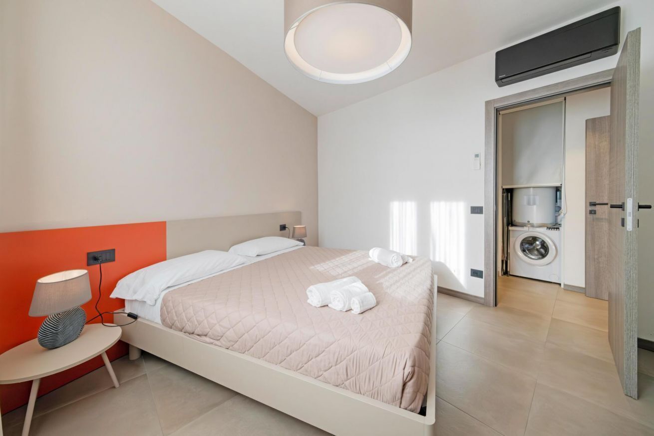 A modern hotel room with a bed, a nightstand, a lamp, a washing machine, and a ceiling light.
