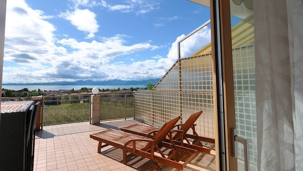 A balcony with a view of the sea and mountains, a tiled floor, and wooden chairs. - ApartmentsGarda - Rosa Chiarè (Bardolino)