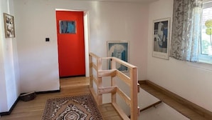 A room with a wooden floor, a red door, and a staircase with a wooden railing.