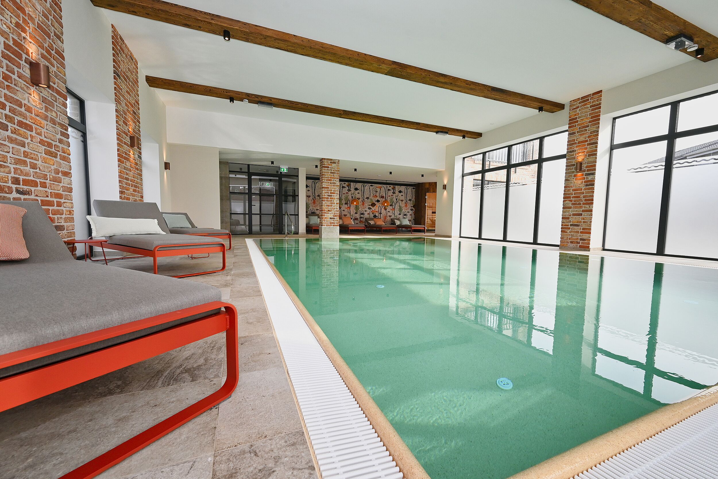 An indoor swimming pool with a modern design, featuring a red-framed lounge area and large windows.