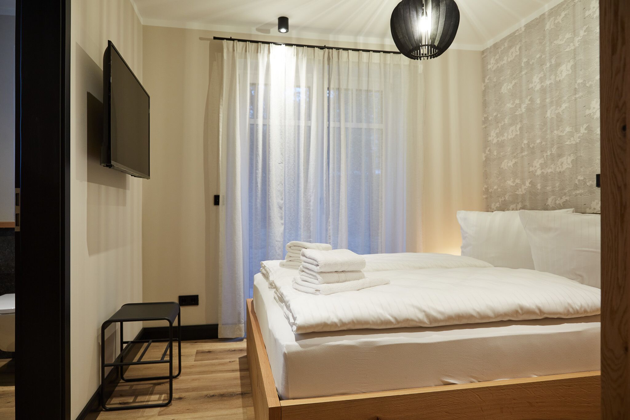 Quarto com cama, televisão na parede e janela com cortinas leves.