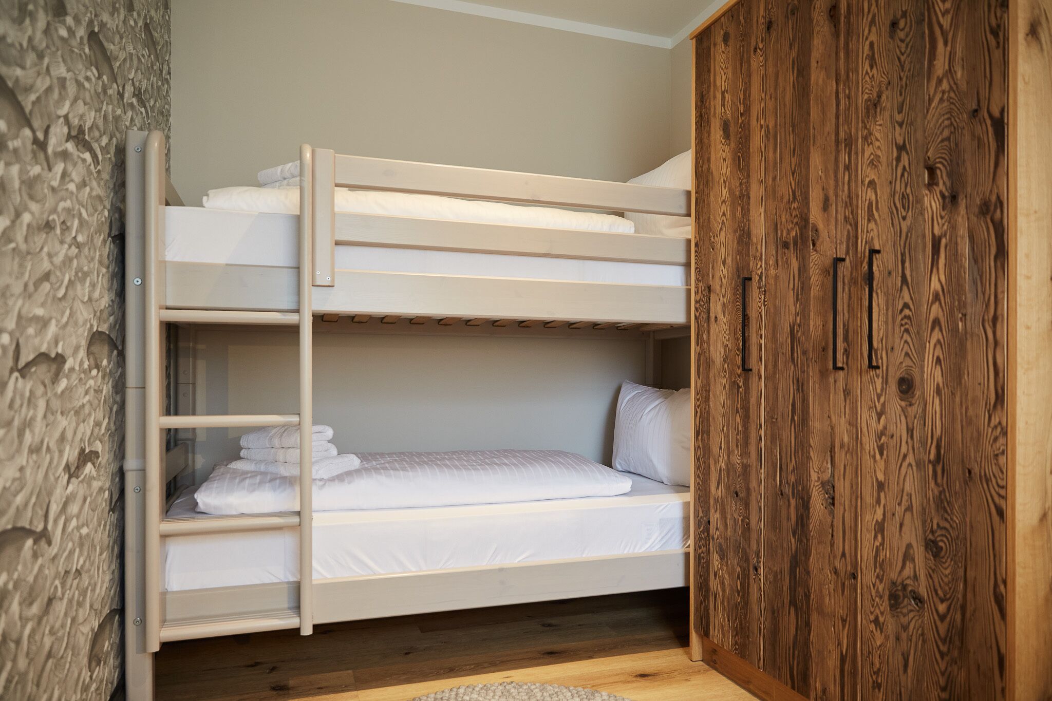A bunk bed with white bedding, a wooden wardrobe, and a stone wall.