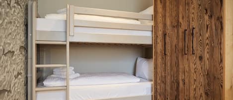 A bunk bed with white bedding, a wooden wardrobe, and a stone wall.
