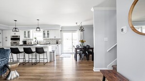 Une cuisine moderne avec des meubles blancs, un îlot central et des tabourets de bar. On y trouve également un coin repas avec une table et des chaises, ainsi qu’un escalier avec une rampe en bois.