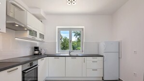 A modern kitchen with white cabinets, a black countertop, a built-in oven, a refrigerator, and a window with a view of trees. - Villa Irena Funtana in a quiet location, with pool close to Zelena Laguna Resort (Funtana)