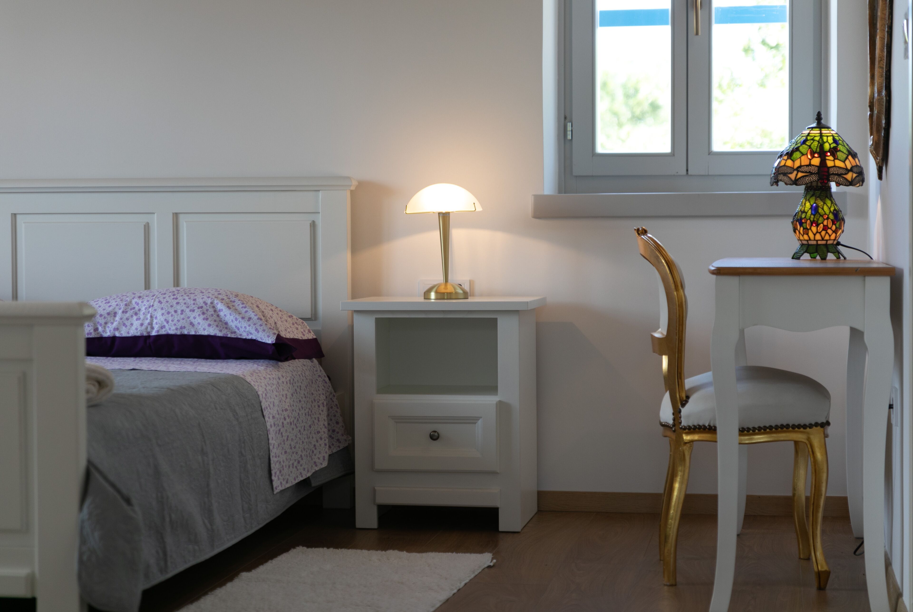 A bedroom with a bed, a nightstand with a lamp, a chair, and a window with a view.