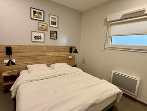 Quarto com cabeceira de madeira, cama com roupa de cama branca, duas mesas de cabeceira com abajures e uma janela com unidade de ar condicionado.