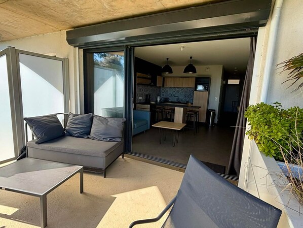 Un espacio habitable moderno con sofá, una mesita, una silla y una cocina visible a través de un amplio ventanal corredero.