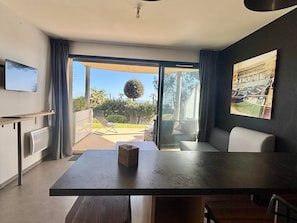 Uma sala de estar moderna com uma mesa grande, um sofá, uma televisão e uma porta de vidro deslizante que dá para uma varanda com móveis de exterior.
