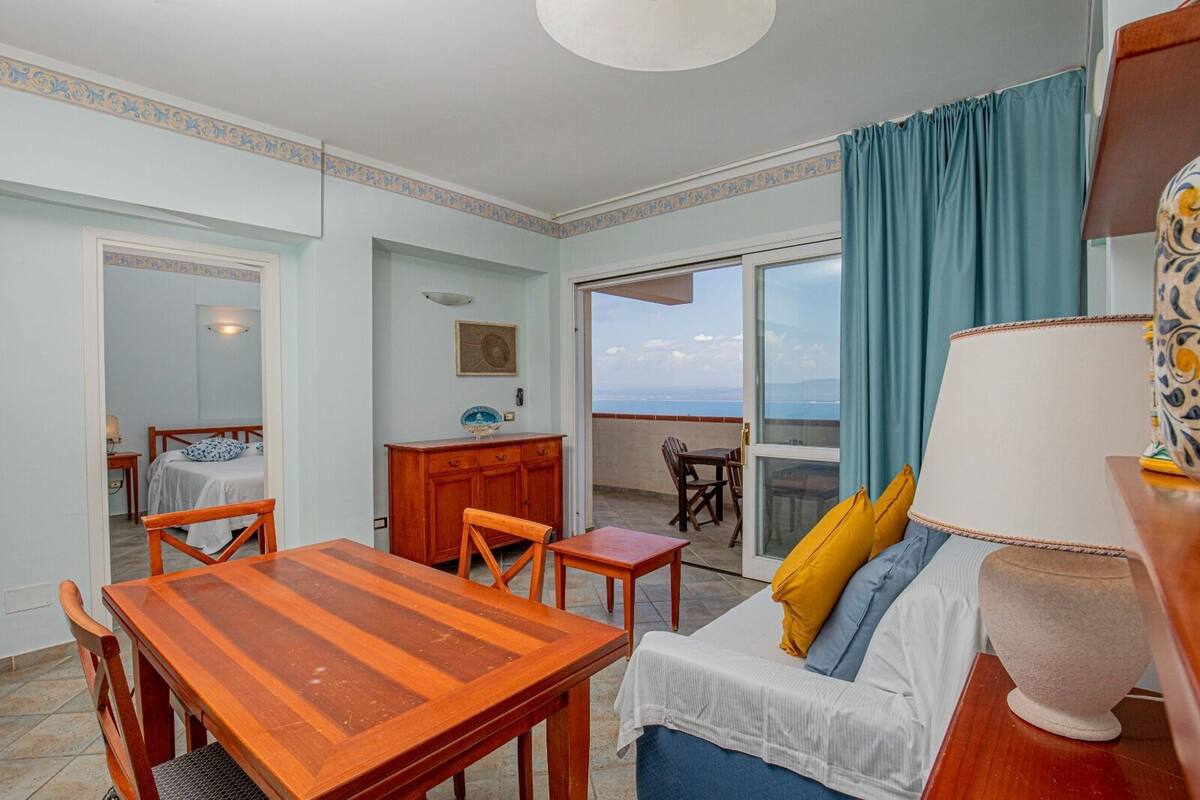 Une pièce avec une table à manger en bois, un canapé avec des coussins, une lampe et une vue sur la mer, visible à travers une porte-fenêtre coulissante.
