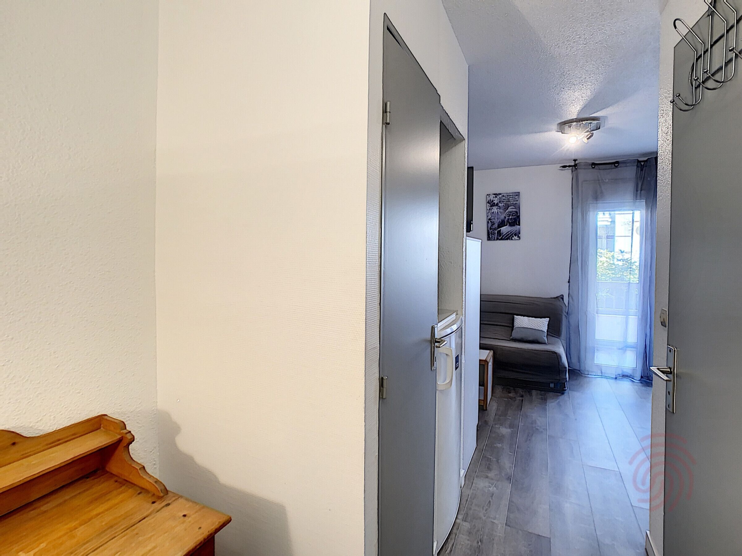 En smal hall som leder till ett rum med en säng, ett litet bord och en dörr.
