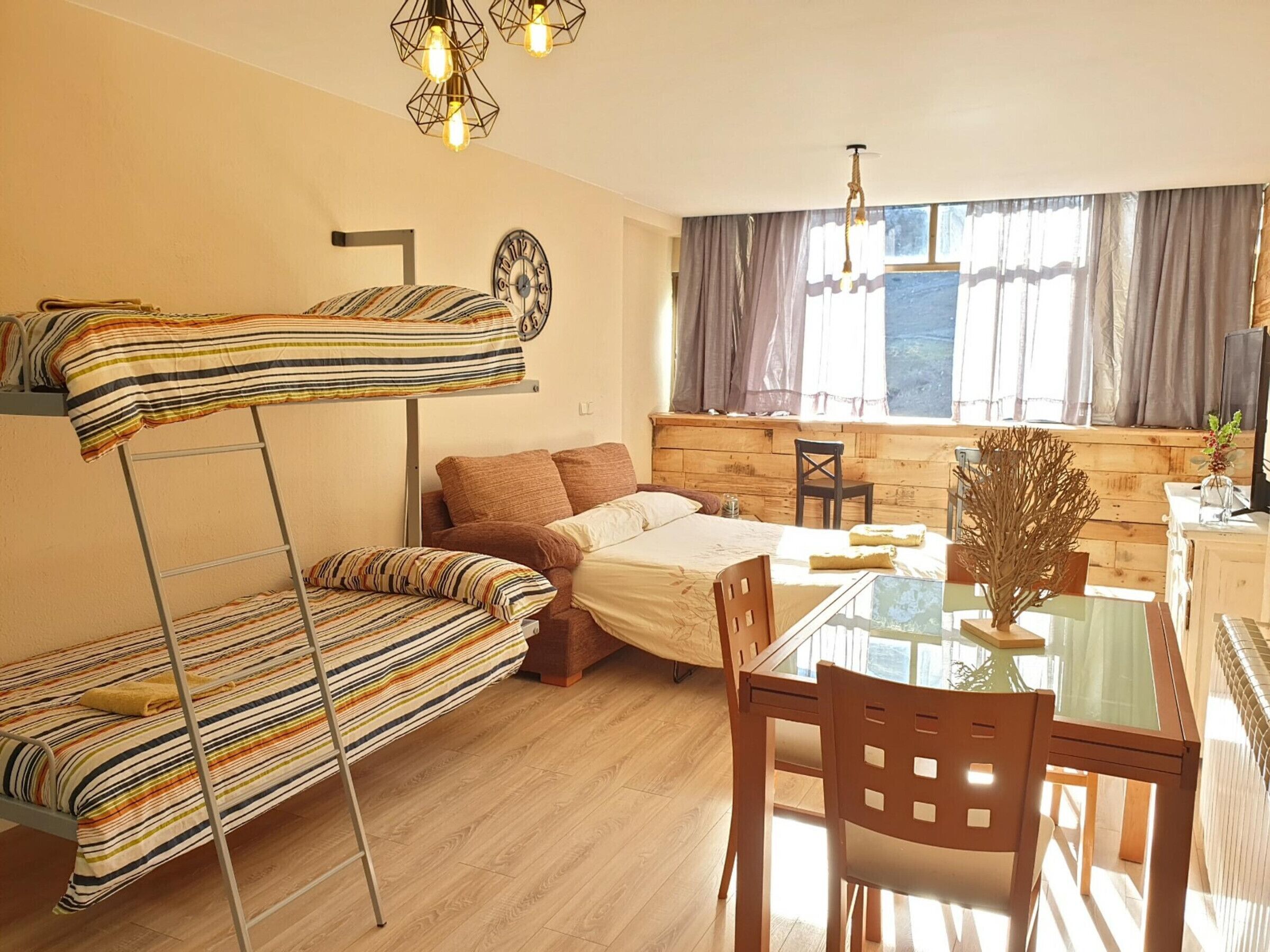 A compact room with a bunk bed, a single bed, a dining table with chairs, a small kitchen area, and a window with curtains.