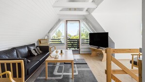 Une chambre de style loft moderne, avec un plafond en pente, un canapé en cuir noir, une table basse en bois, un téléviseur et une grande fenêtre donnant sur un paysage verdoyant.