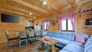 A cozy cabin interior with a wooden ceiling, a dining area with a table and chairs, a blue sofa, and a kitchen area with a refrigerator and a clock.