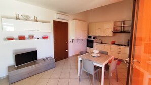A modern apartment with a living area featuring a flat-screen TV, a sofa, and a coffee table. There is a dining area with a table and chairs, and a kitchen with cabinets and appliances.