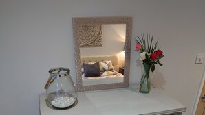 Une commode blanche avec des tiroirs, un miroir avec un cadre décoratif, un vase en verre avec des fleurs et un bocal en verre rempli de perles.