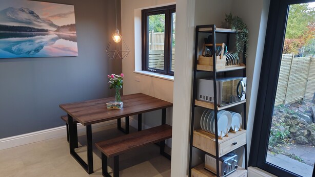 Un coin repas avec une table en bois, un banc et une étagère en métal où sont rangés de la vaisselle et des ustensiles de cuisine.