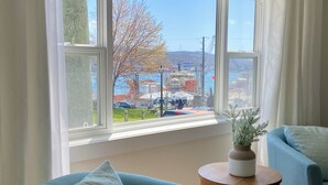 A room with a large window, a blue armchair, a small wooden table with a plant, and a view of a park and buildings outside. - Signal Hill Elegance 3Bd A/C Hiking (St. John's)