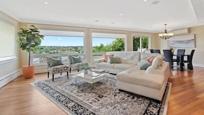 A spacious living room with a large window, a white sofa, a zebra-patterned chair, a coffee table, and a dining area with a chandelier.