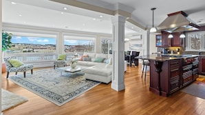 A spacious living room with a large white sofa, a coffee table, and a patterned rug. There is a kitchen area with dark wooden cabinets and a large island.