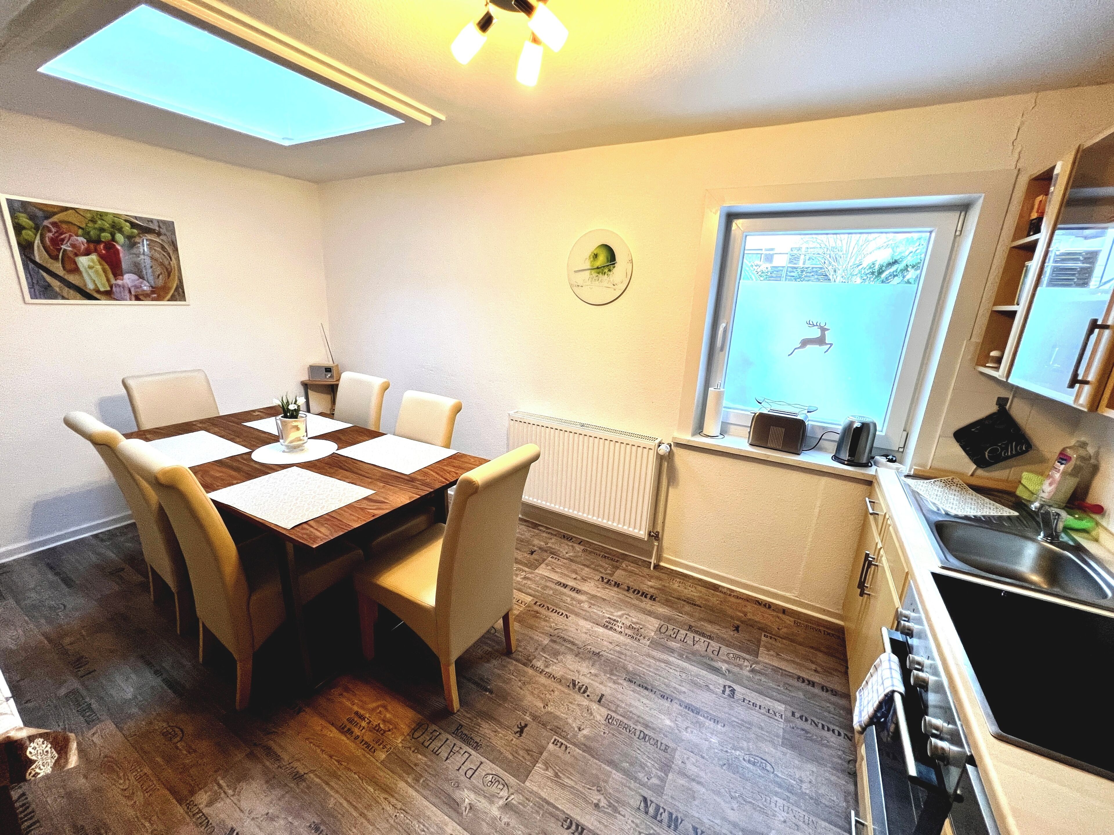 Un comedor con una mesa y sillas de madera, un radiador, una ventana con vista a un ciervo y una cocina con un lavaplatos y estufa.