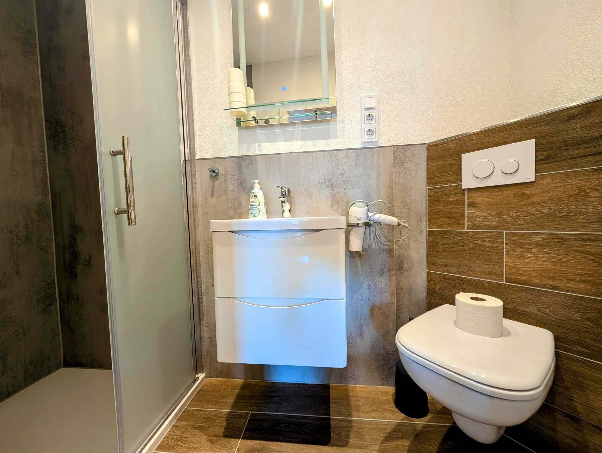 Een moderne badkamer met een douchecabine van glas, een wit wastafelmeubel met een glazen plank erboven en een toilet met een rol toiletpapier ernaast.