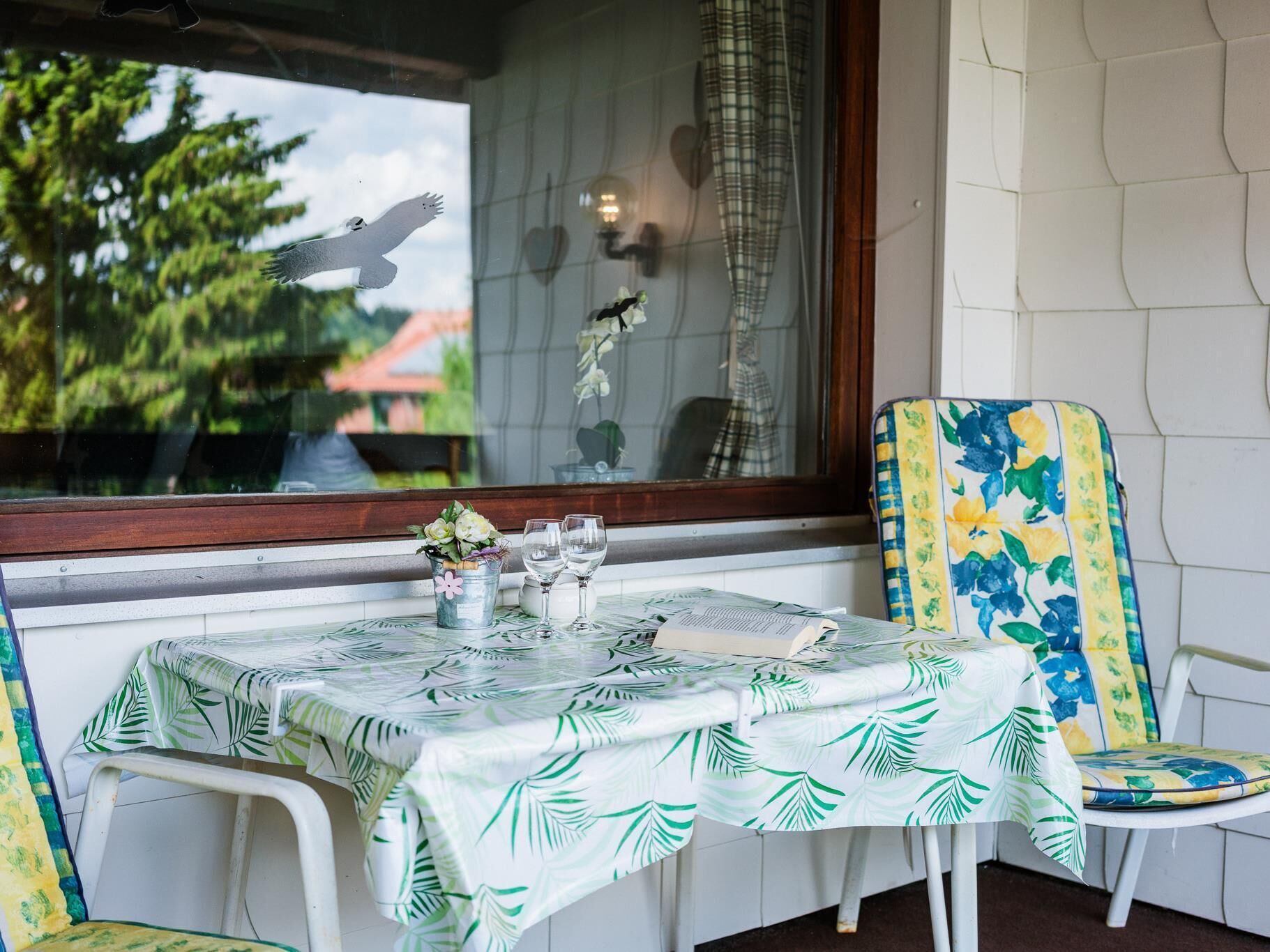 Uma mesa posta com toalha de folhagem tropical, uma cadeira com padrão floral, e uma janela com vista para árvores e um edifício.