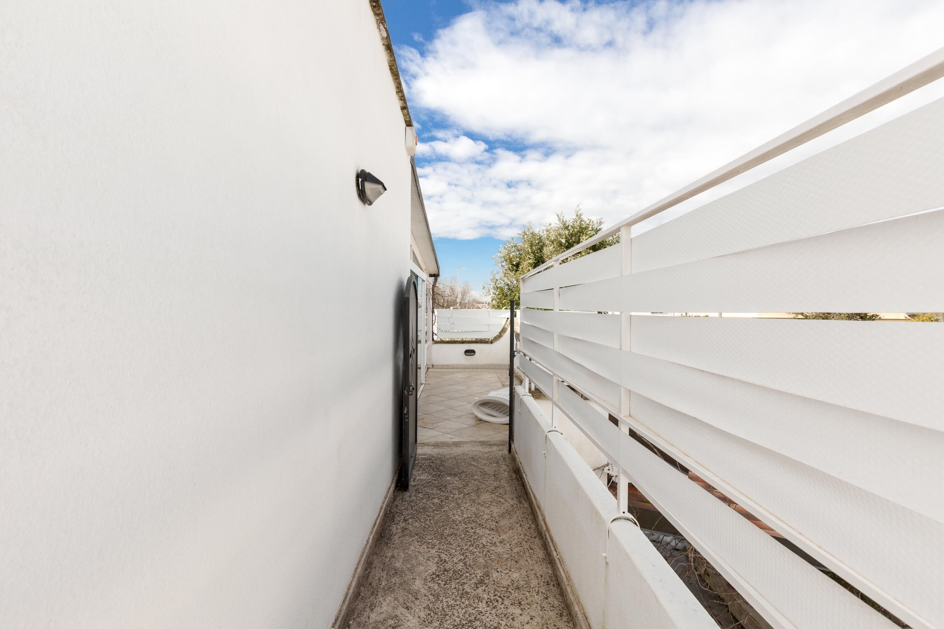 A narrow corridor with white walls and a concrete floor, leading to a white fence and a building in the distance.