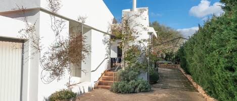 Un bâtiment blanc avec un escalier menant à une porte, entouré de verdure et bordé d’un chemin pavé.