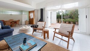 A spacious living room with a blue sofa, wicker chairs, a coffee table with magazines, and a large sliding glass door leading to an outdoor patio with potted plants.