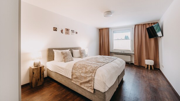 Quarto com cama grande, mesa de cabeceira, televisão de tela plana na parede e janela com cortinas.
