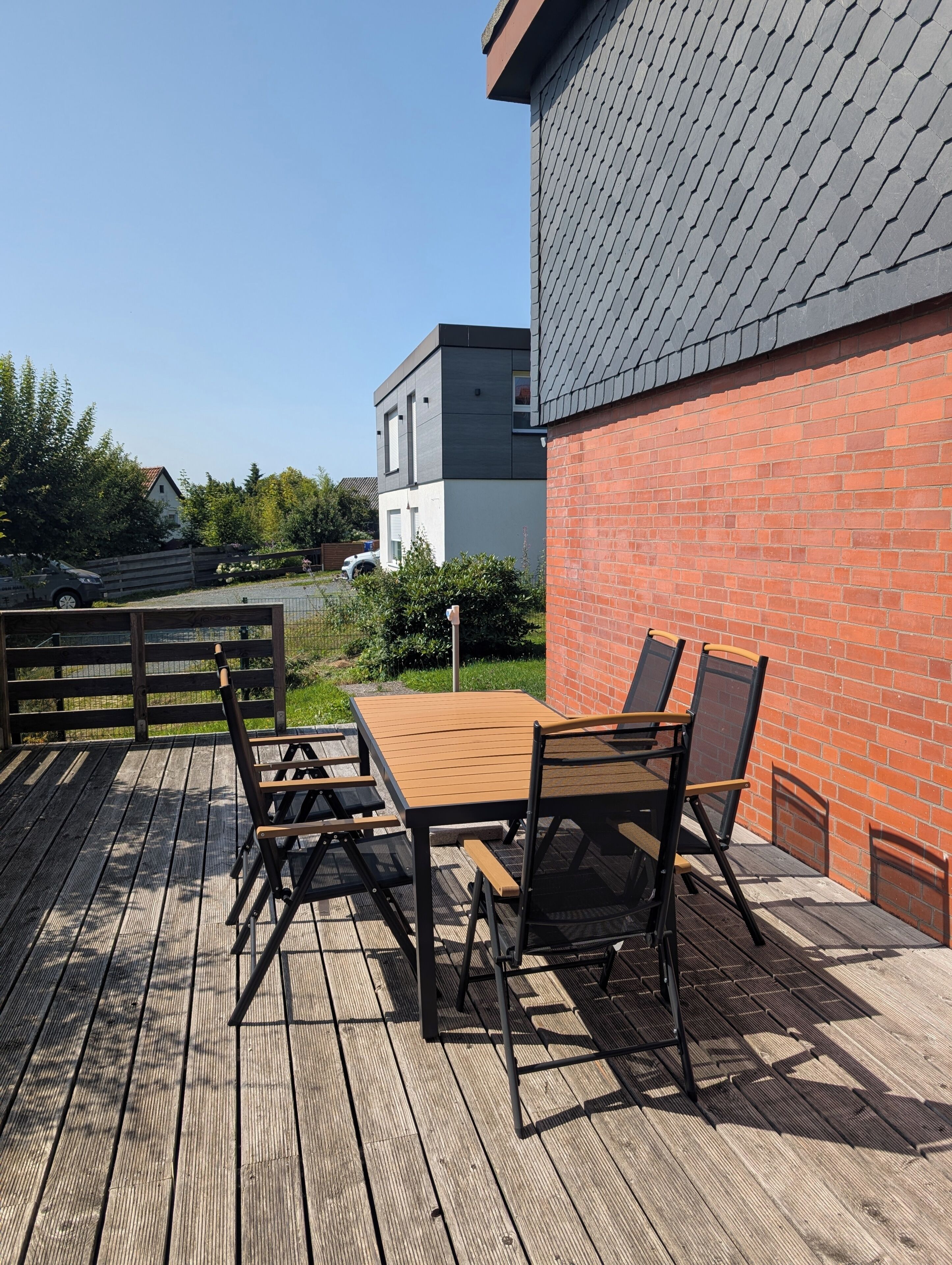 Eine Terrasse im Freien mit einem Holztisch und -stühlen, einer Ziegelwand und einem modernen Gebäude im Hintergrund.