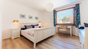Quarto com cama, mesas de cabeceira, escrivaninha, cadeira e uma janela grande com cortinas.