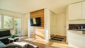 A modern living space with a sofa, a small table with a vase, a flat-screen TV mounted on a wooden wall, and a kitchen area with white cabinets and a countertop. - Holiday Apartment "Fichte" (Braunlage)