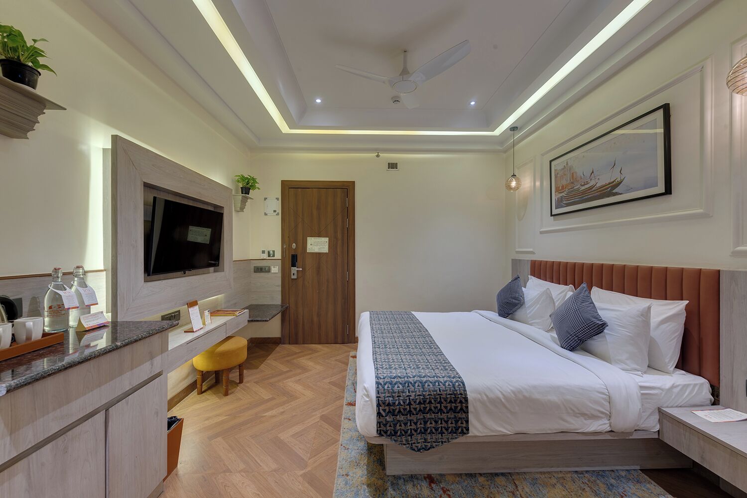 A hotel room with a bed, bedside table, TV, desk, chair, and a painting on the wall.