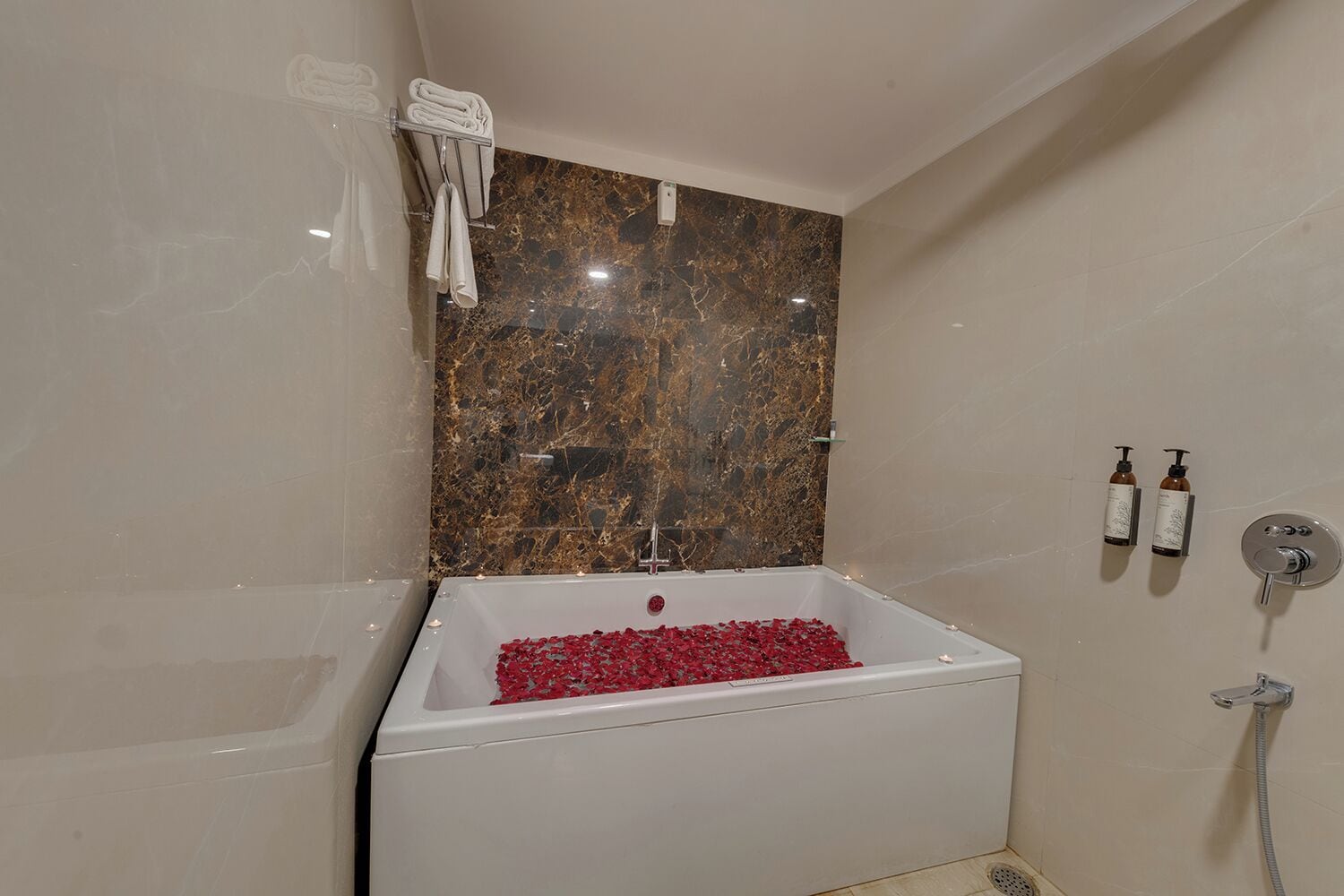A bathroom with a marble wall, a bathtub filled with rose petals, and two bottles of shampoo.