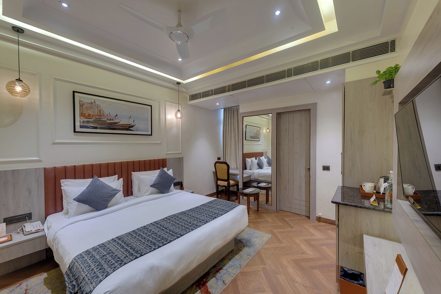 Quarto de hotel com uma cama grande, mesas de cabeceira, escrivaninha, cadeira, espelho e planta em vaso.
