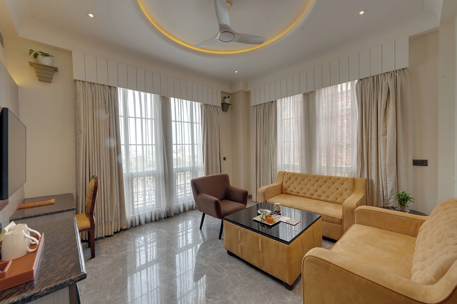 A modern living room with a sofa, armchair, and coffee table, large windows with curtains, a ceiling fan, and a flat-screen TV.