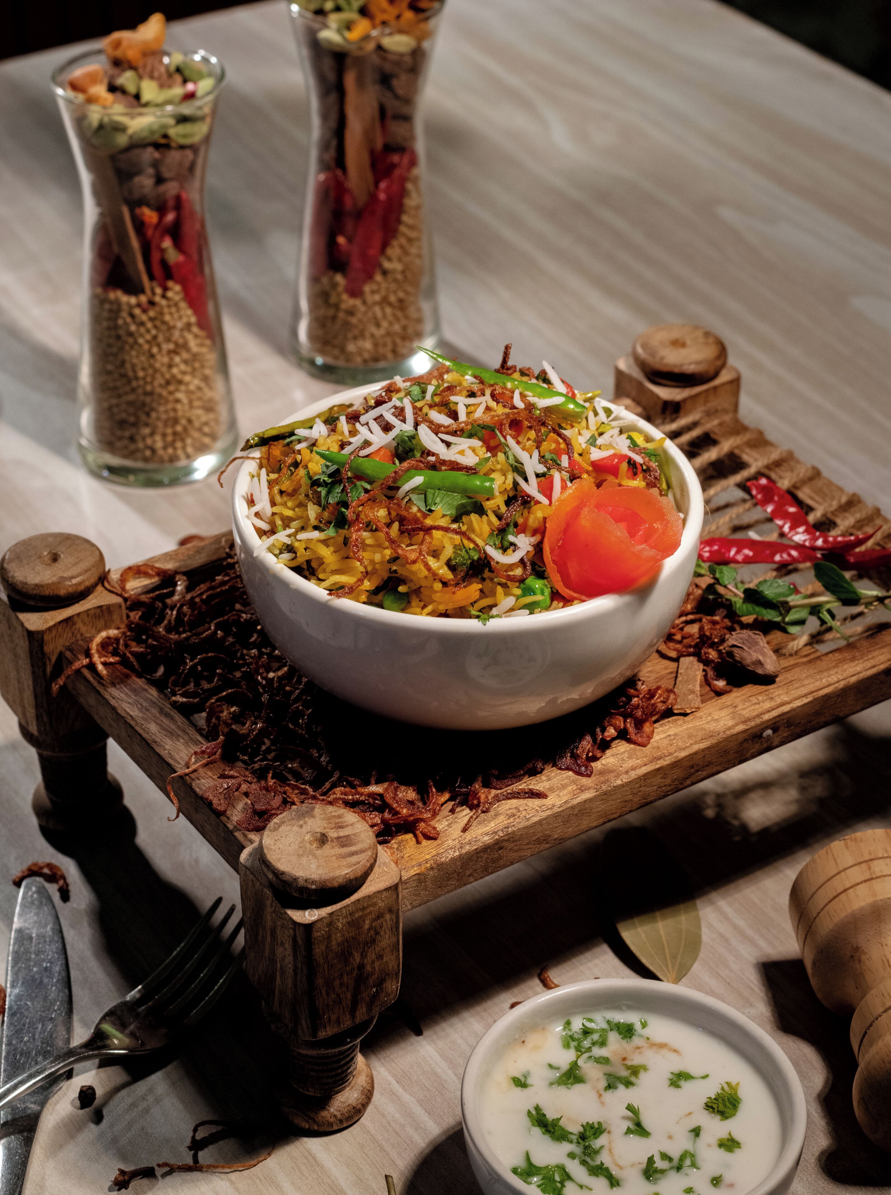 A bowl of rice dish with vegetables and herbs, a small bowl of sauce, and a wooden serving board with a fork and spices.