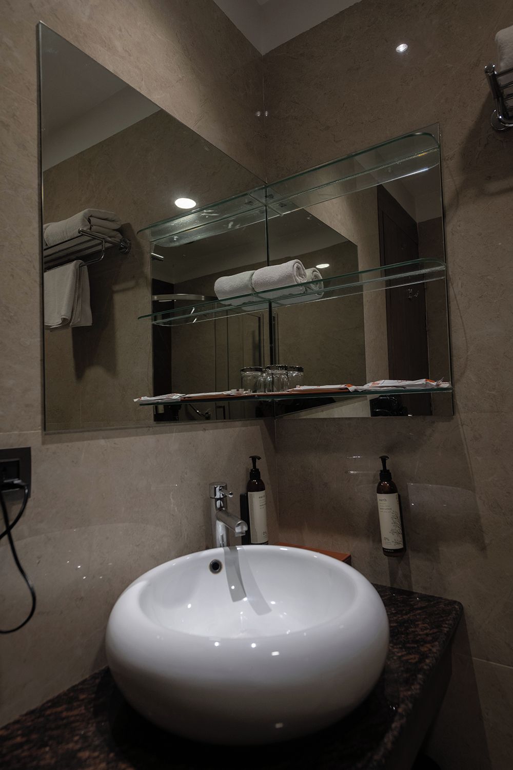 A bathroom with a round white sink, a glass shelf above the sink, and a mirror reflecting a towel and a glass jar.