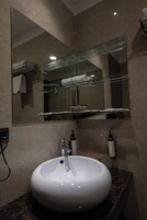 Een badkamer met een rond wit wastafeltje, een glazen plank erboven en een spiegel die een handdoek en een glazen pot weerspiegelt.