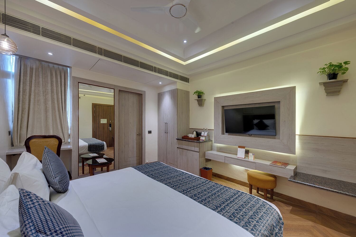 A modern hotel room with a large bed, a desk with a chair, a TV mounted on the wall, and a small plant.