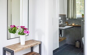 Une salle de bain avec un lavabo blanc, des carreaux foncés et un banc en bois sur lequel est posé un vase de fleurs.