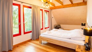 Een slaapkamer met een houten plafond, een bed met witte lakens, een nachtkastje met een lamp, en uitzicht op groen door grote ramen met rode frames.