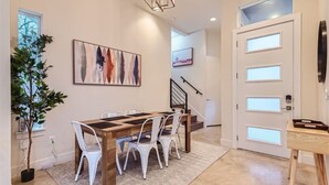 Uma sala de jantar com mesa de madeira e cadeiras brancas, um quadro na parede, uma planta em vaso e uma escada que leva a outro cômodo.