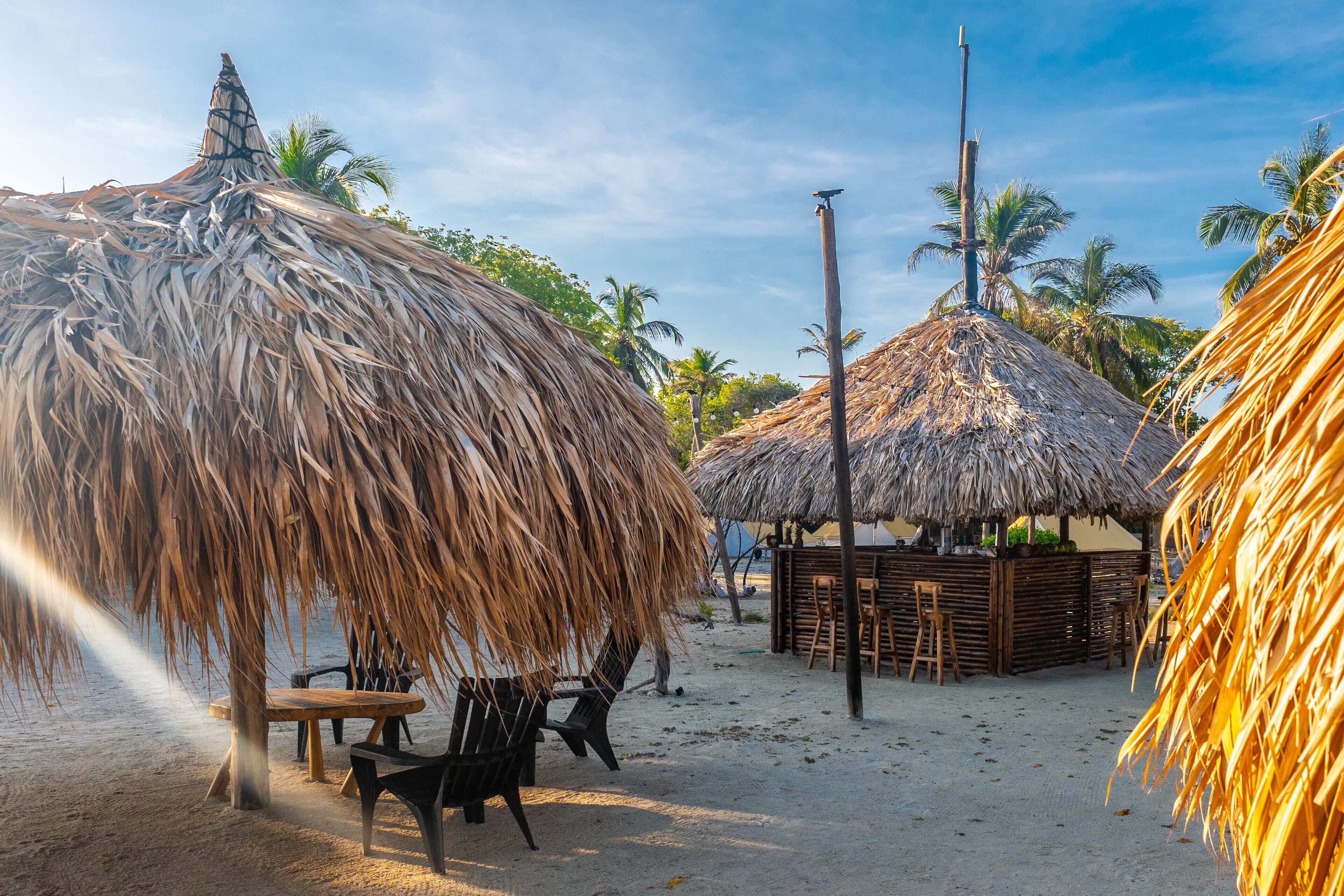 A beach scene with thatched-roof huts, wooden furniture, and palm trees.